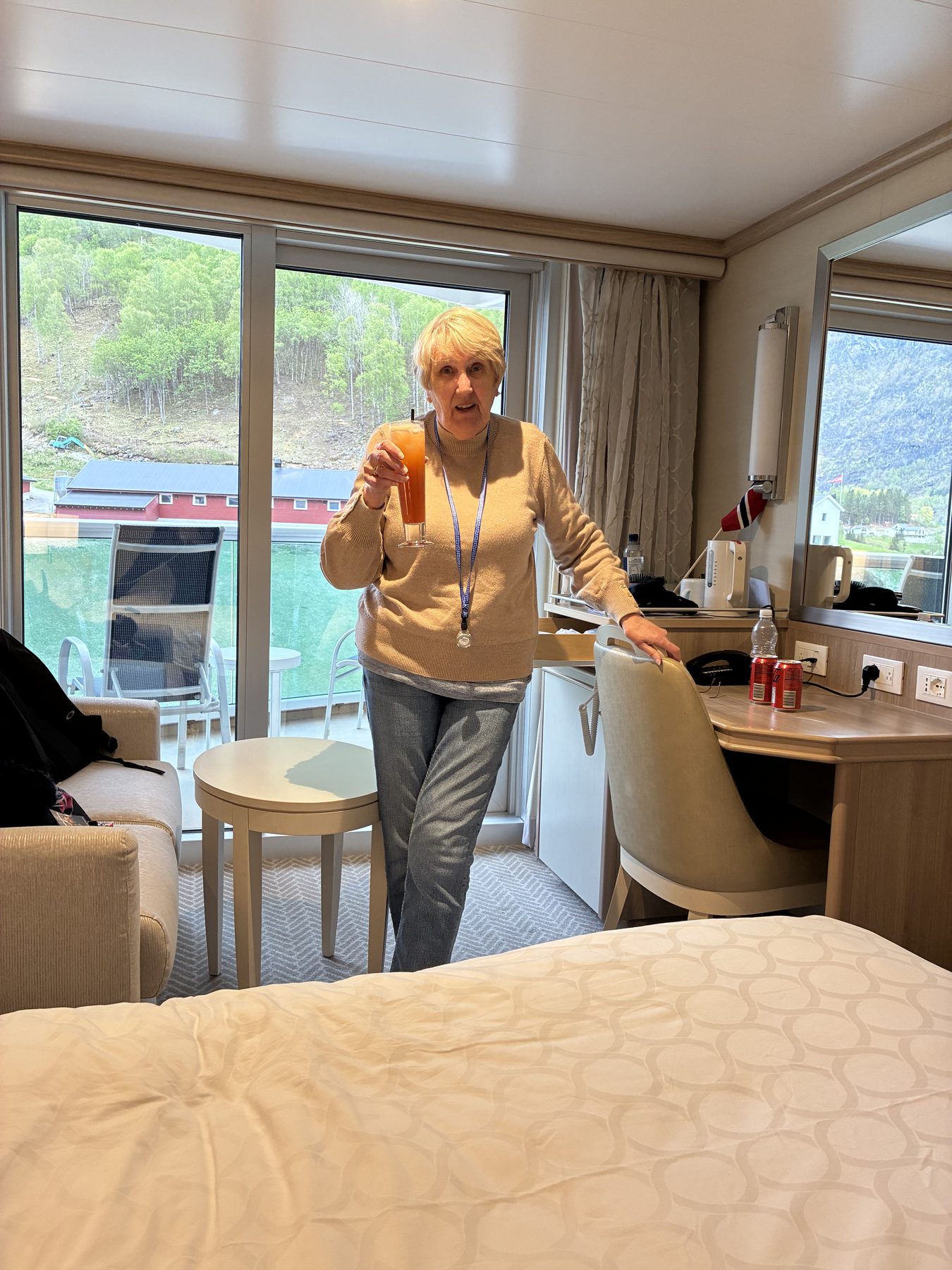 Frances holding an orange cocktail in the cabin, fjord visible through the balcony doors