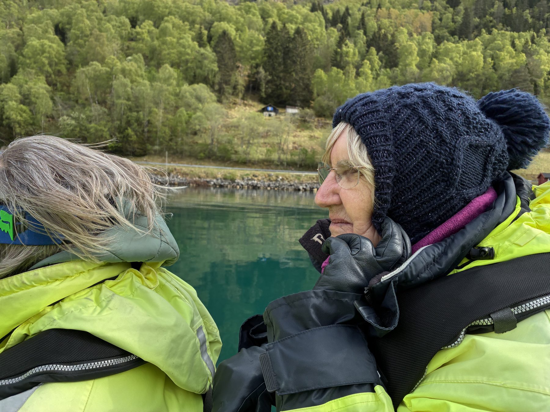 Frances looking out across the green fjord water, navy bobble hat, gloves on